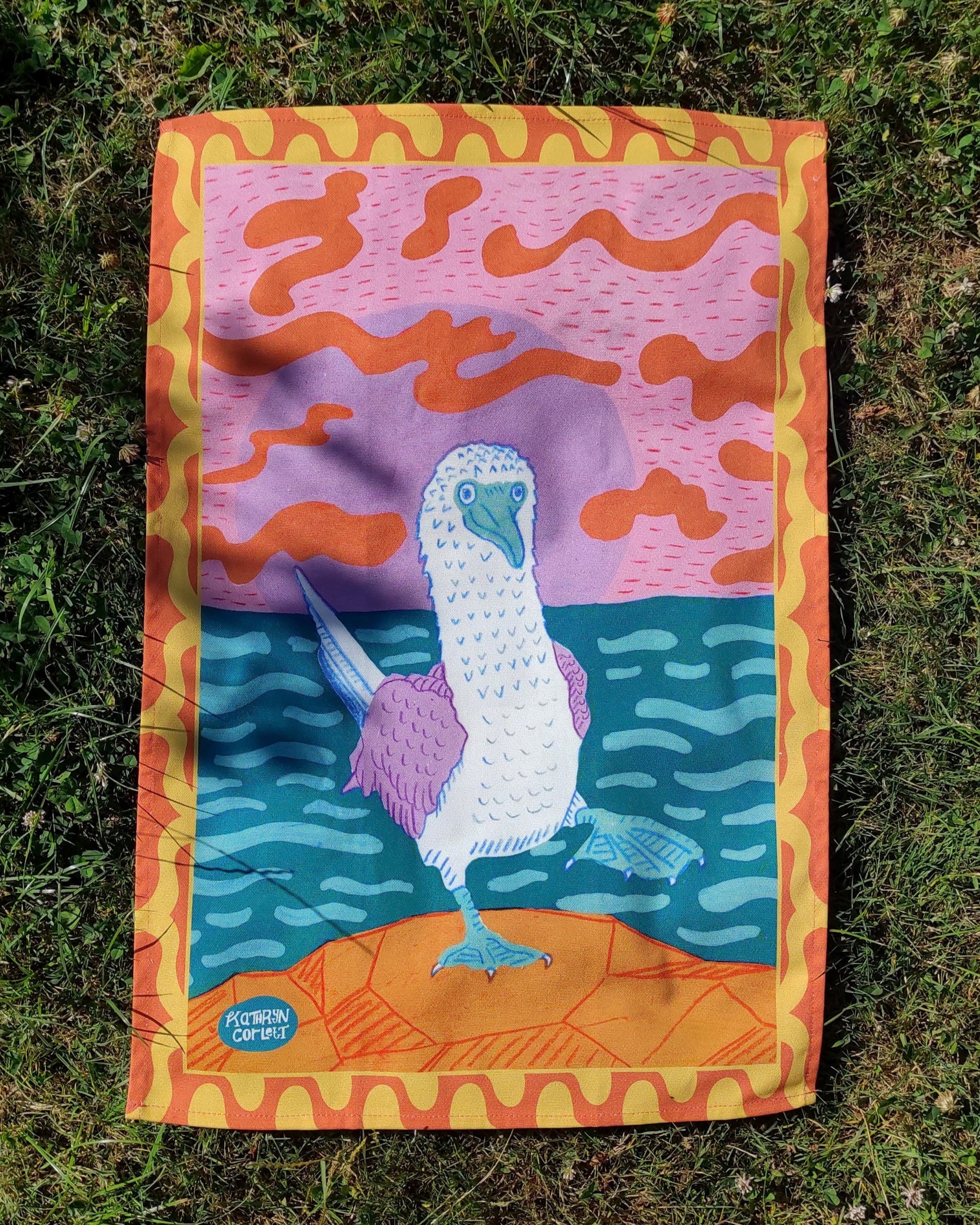 A colourful tea towel with a Blue Footed Booby design on grass
