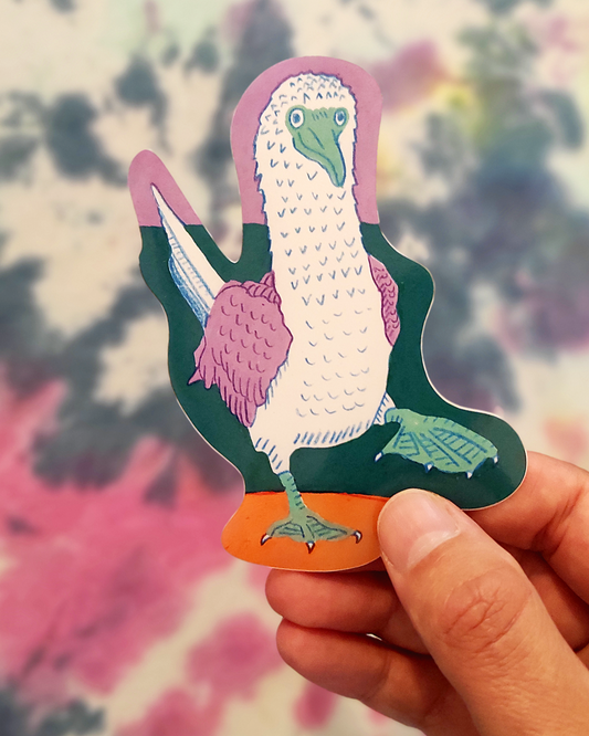 Hand holding a colorful sticker of a blue footed booby bird with a blurred nature background