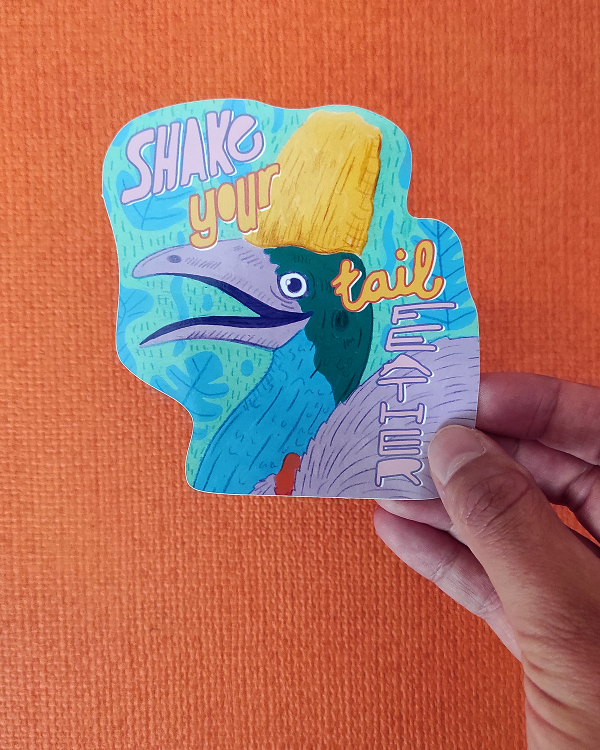 Colorful card with a bird design and text 'Shake your tail' held against an orange background