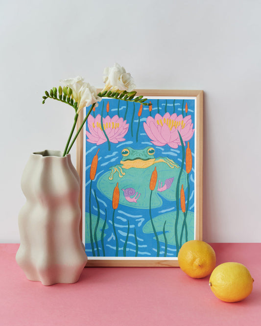 Framed artwork of a frog and lilies on a pink surface with a vase and lemons.