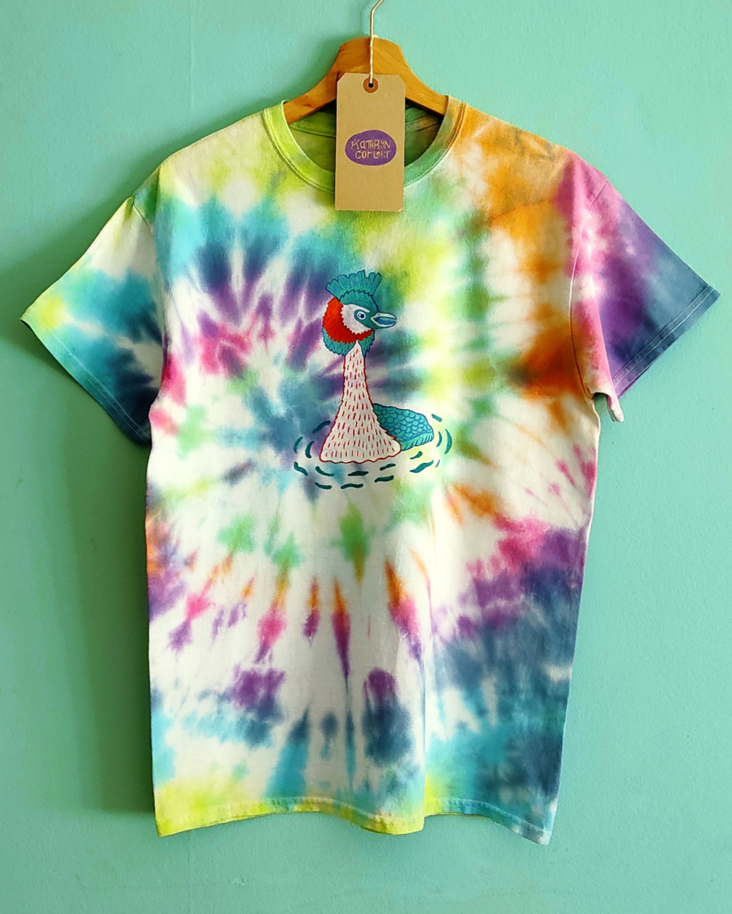 Colorful tie-dye t-shirt with a graphic design or a Great Crested Grebe on a hanger against a green background