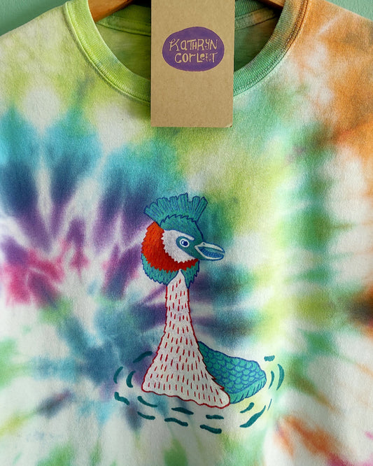 Tie-dye t-shirt with a great crested grebe design and a tag on a hanger against a green wall.