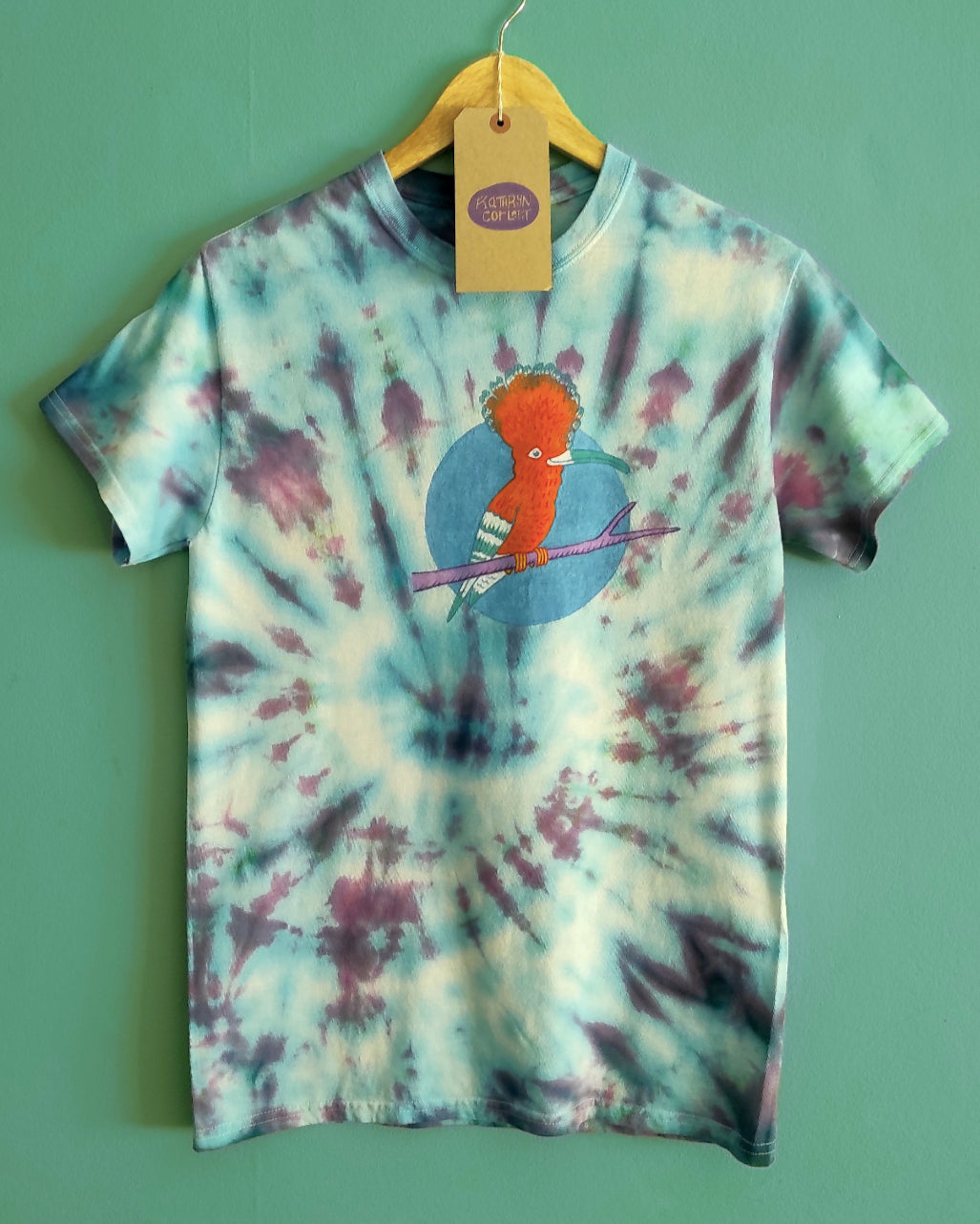 Tie-dye t-shirt with Hoopoe illustration on a hanger against a green background