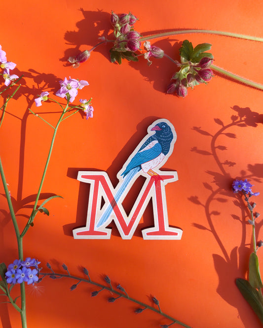 Letter 'M' with a Magpie design on an orange background with floral elements