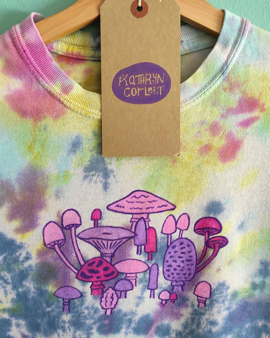Tie-dye shirt with mushroom design and a tag on a hanger against a light green wall.