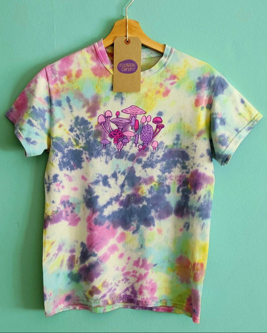 Colorful tie-dye t-shirt with hand drawn mushrooms in pink and purple on the front on a hanger against a green wall