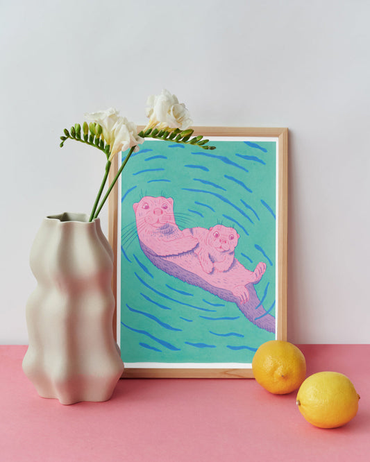 Framed artwork of an otter Mum and Pup floating in water with a vase, flowers, and lemons on a pink surface.