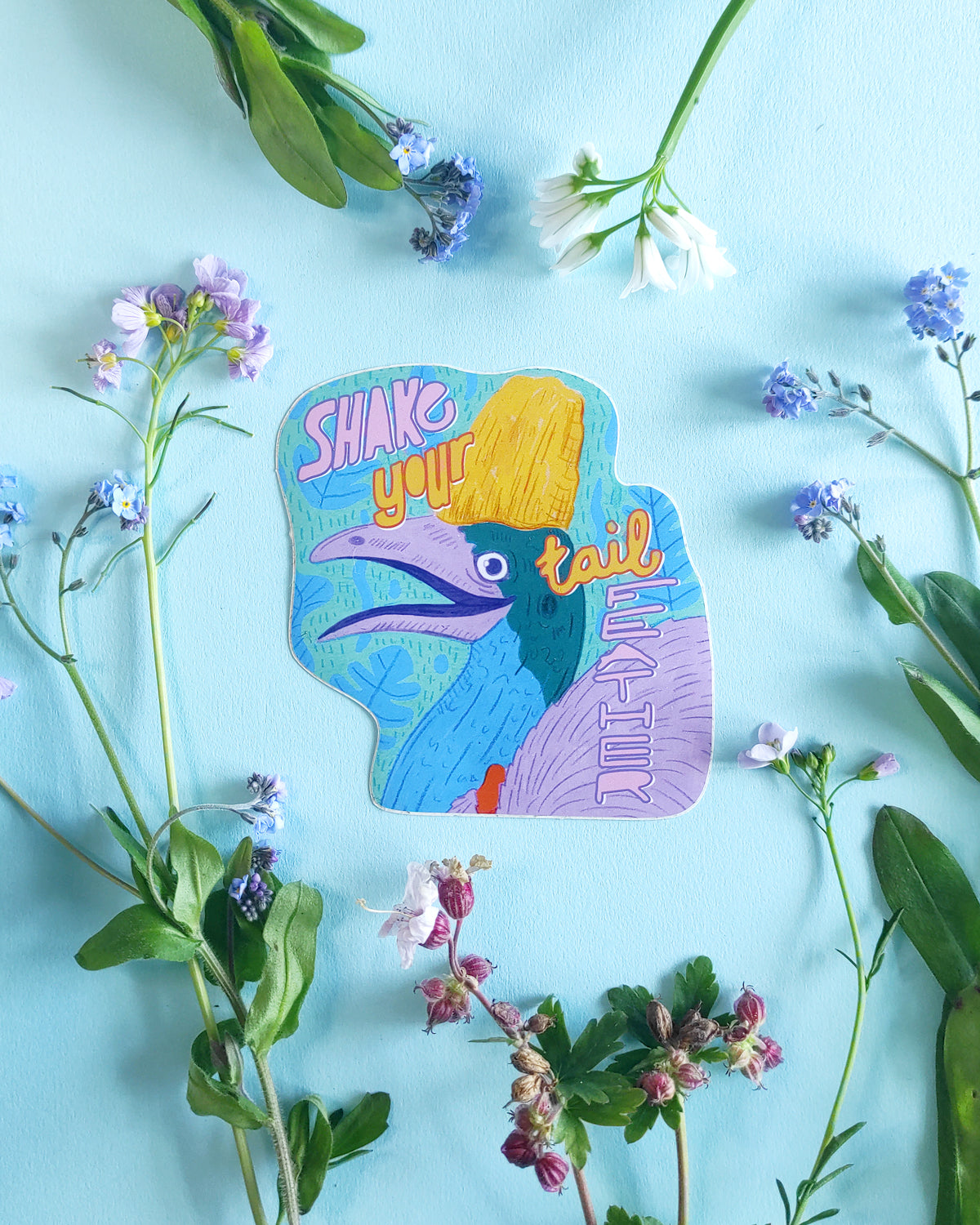 Colorful sticker with a bird design and text 'Shake your tail' surrounded by flowers on a light blue background