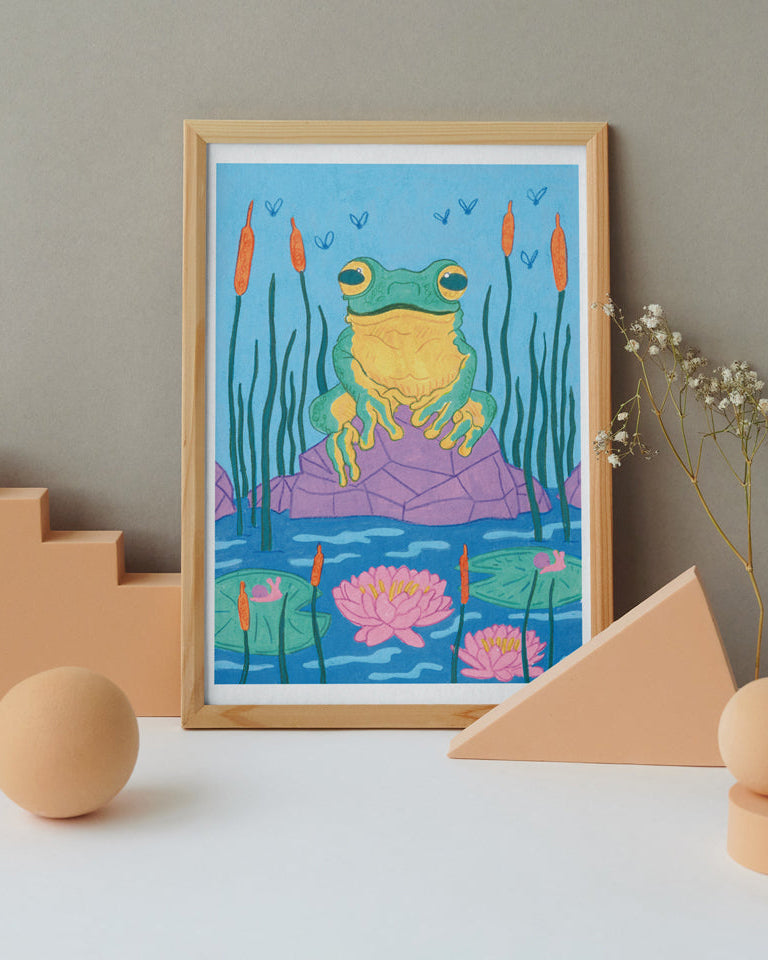 Framed artwork of a frog in a lily pad pond with geometric objects on a white surface.