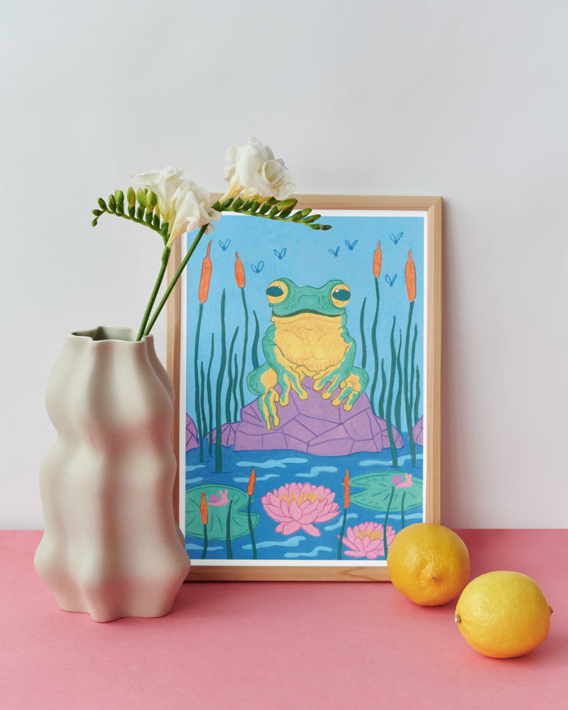 Framed artwork of a frog in a pond with lilies and reeds, placed on a pink surface with a vase and lemons.