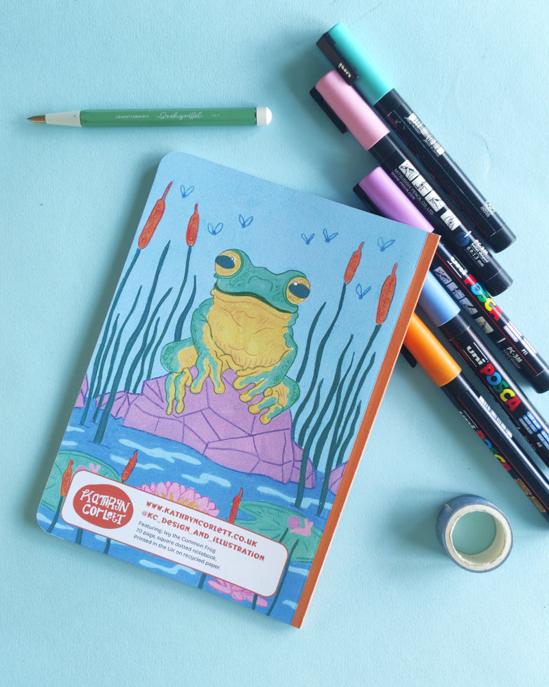 Notebook with frog design on a blue background with markers and tape