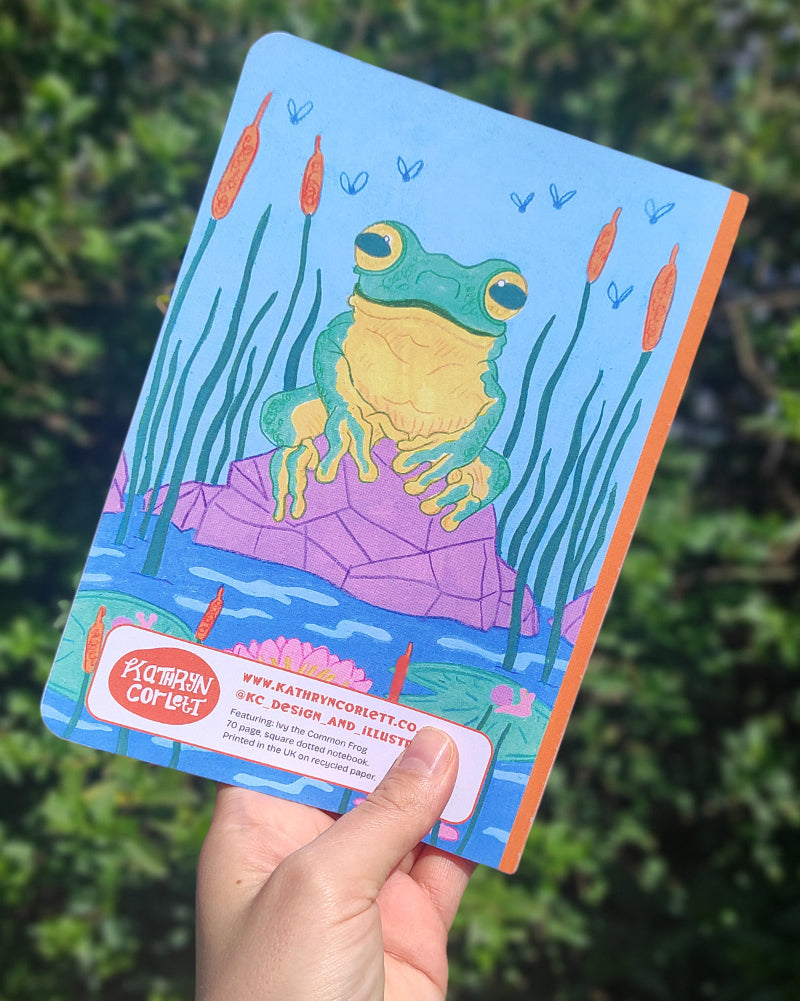 Colorful notebook with a frog illustration held by a hand against a green leafy background