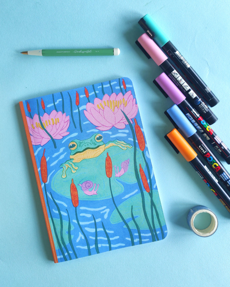 Notebook with a frog and water lily design on a blue background with markers and tape.