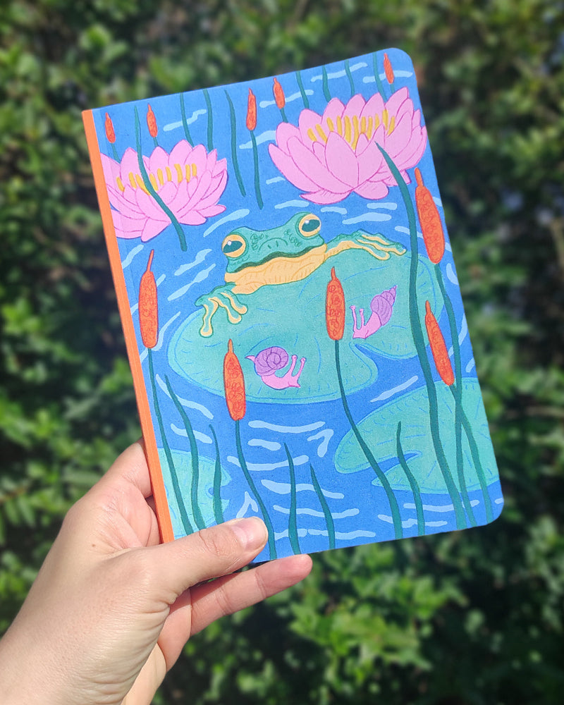 Notebook with a frog and lily pad design held in front of green foliage