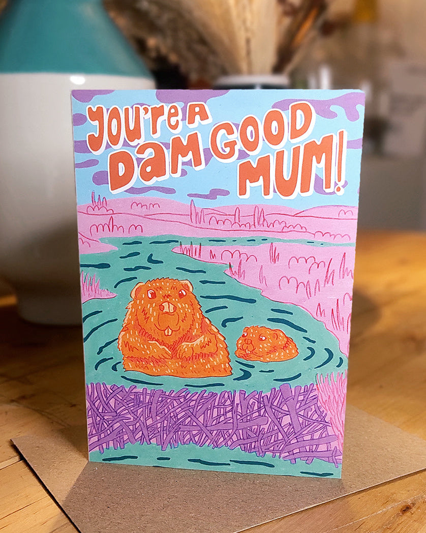 Greeting card with beaver and kit illustration and text 'You're a dam good mum!' on a table