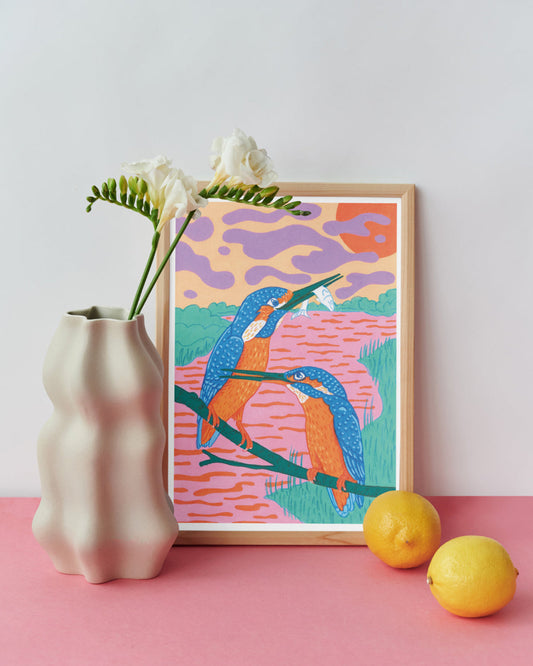 Colorful kingfisher illustration in a wooden frame on a pink surface with a vase and lemons.