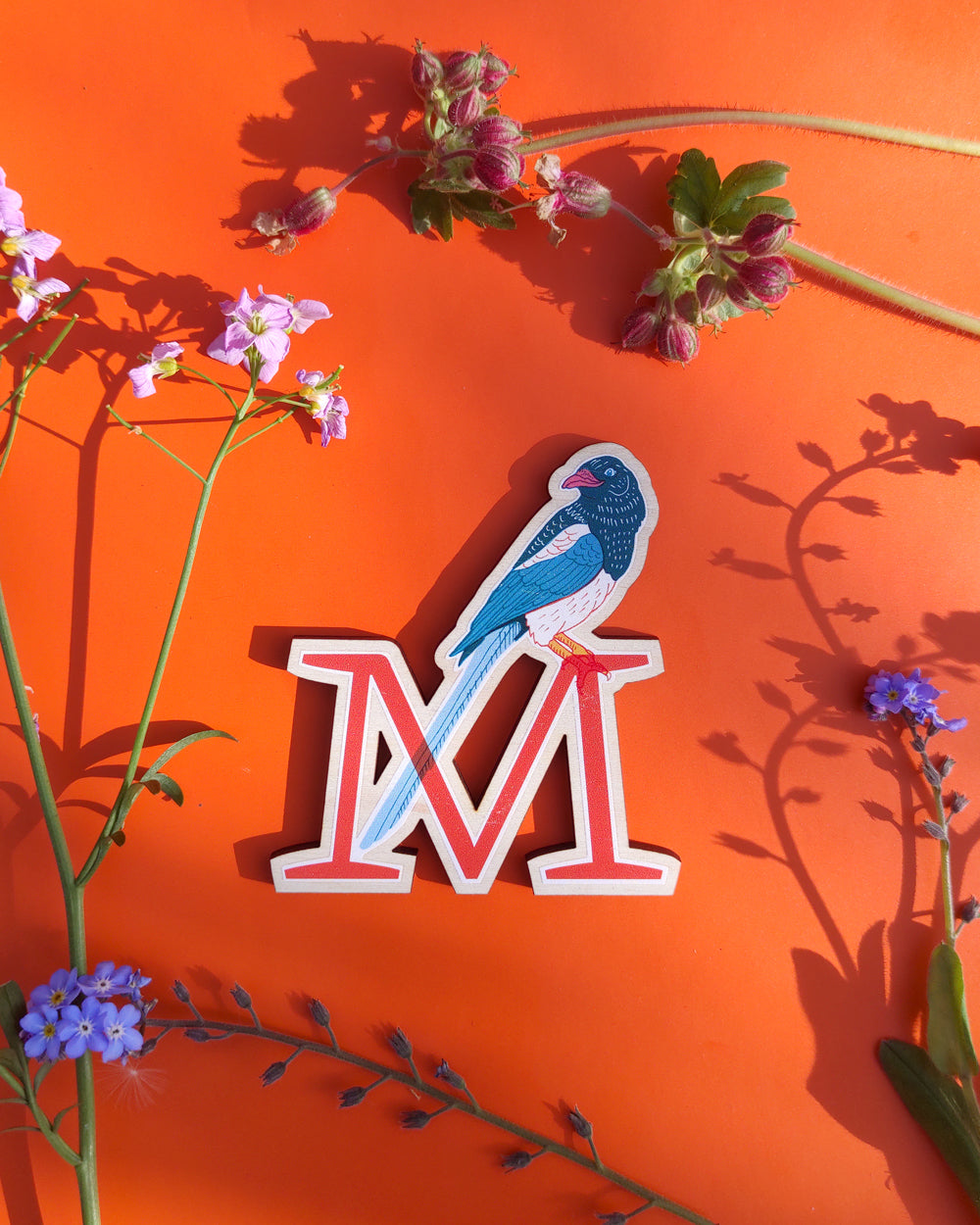 Letter 'M' with a Magpie design on an orange background with floral elements
